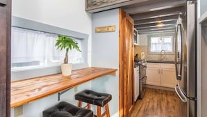 Una cocina compacta con encimera de madera, dos taburetes, un refrigerador y una ventana con una planta.
