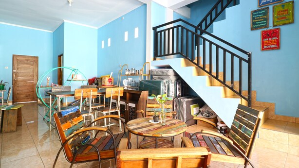 A room with a blue wall, a wooden door, and a staircase with black railings. There are several chairs and tables, some with colorful patterns. - Balini 9 Hostel (Denpasar)