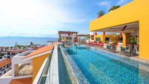 Una piscina infinita en la azotea con vistas al mar, rodeada de edificios amarillos y sillones de baño rojos.