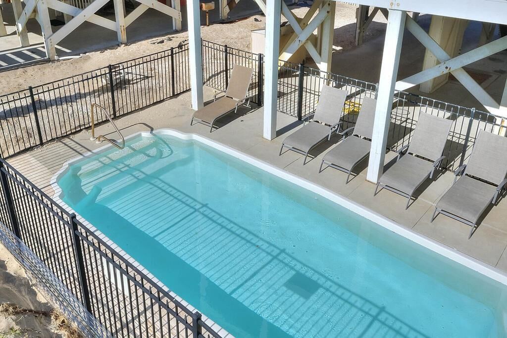 Una piscina rectangular amb superfície d'aigua blava i cristal·lina, envoltada per una tanca de metall negre. Diversos relaxants estan alineats al costat de la piscina, i hi ha una estructura blanca amb teulada.