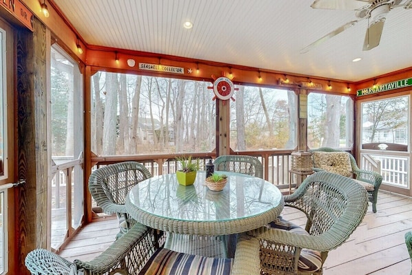 A covered outdoor deck with wicker furniture, a glass table, and a view of trees.