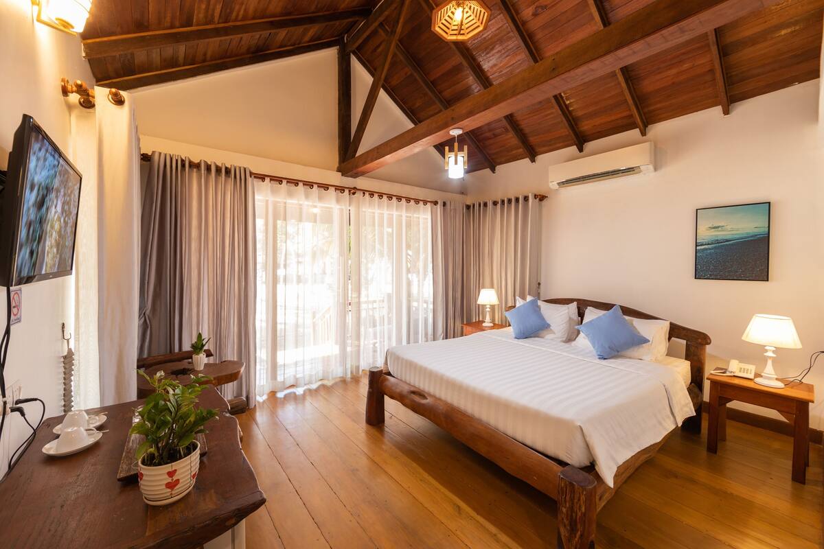 A bedroom with a wooden bed, a bedside table with a lamp, a potted plant, and a view of the outdoors through a window.