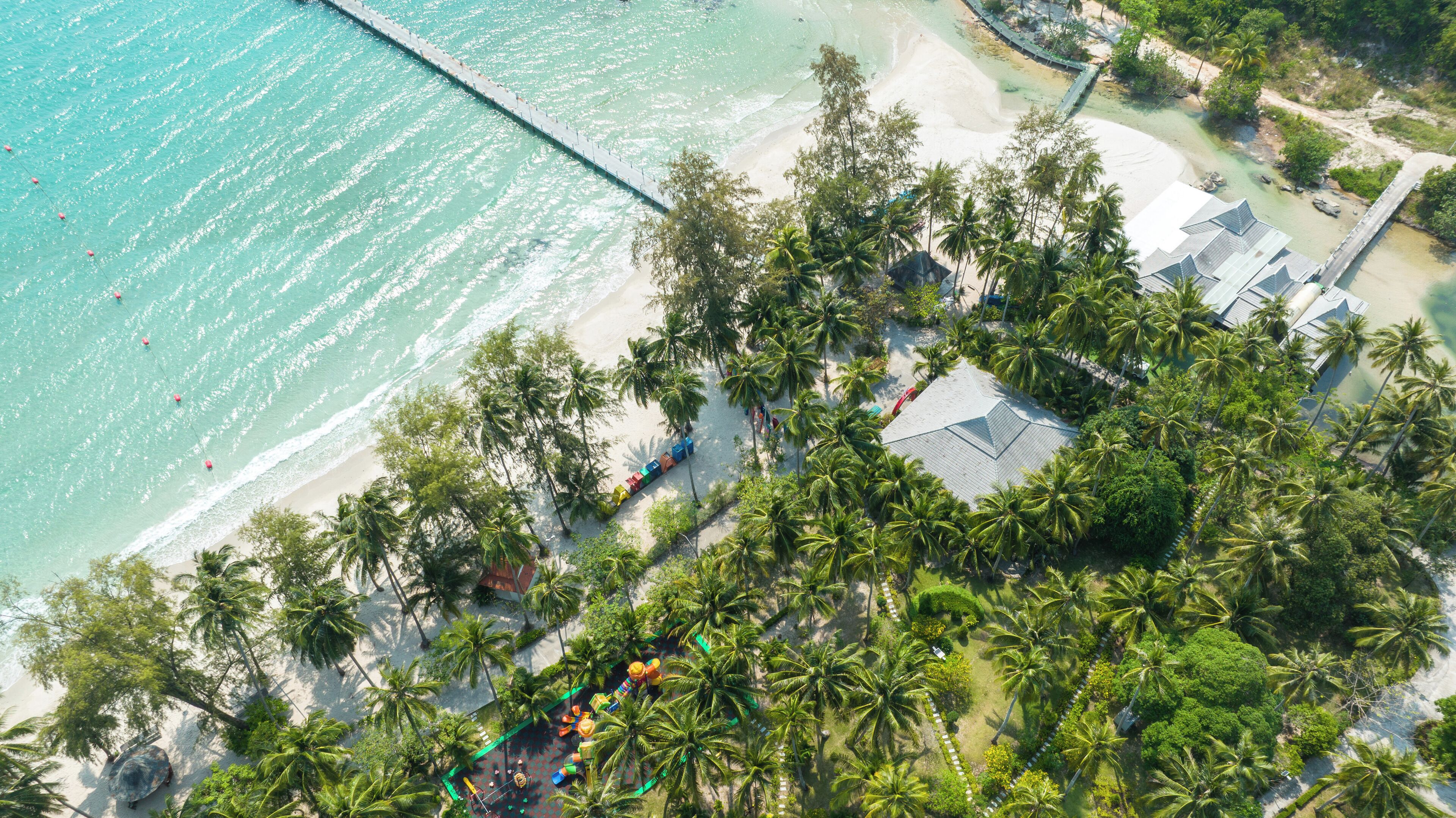 Vista aérea de um resort à beira-mar, com um longo píer, palmeiras e um pequeno edifício com telhado inclinado.