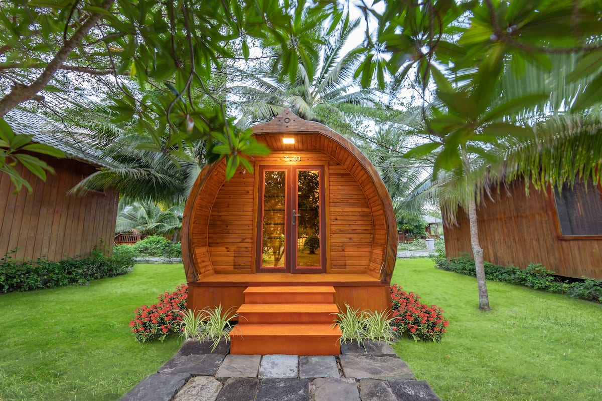 A wooden cabin with a curved roof, surrounded by lush greenery and palm trees.