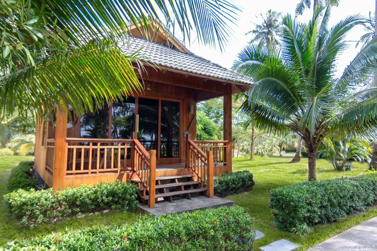 A wooden cabin with a porch, surrounded by lush greenery and palm trees.