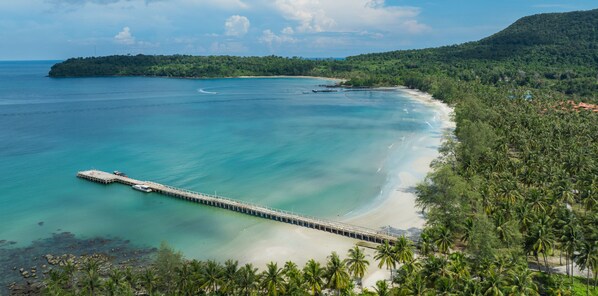 A coastal area with a pier extending into turquoise waters, surrounded by lush greenery and a distant hill. - Mafiya Resort (Koh Rong)