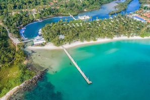 An aerial view of a coastal resort with a long pier extending into turquoise waters, surrounded by lush greenery and a sandy beach.
