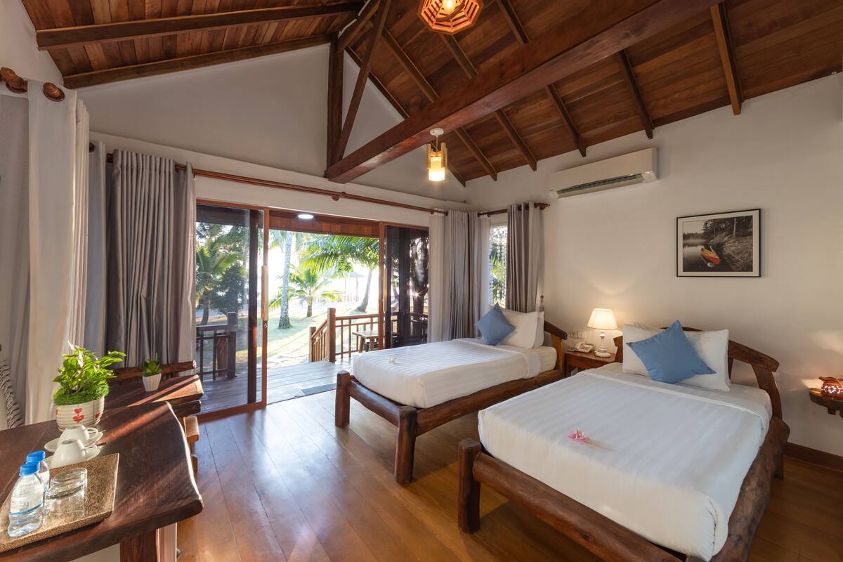 A room with two wooden beds, a wooden desk with a plant, a table with a bottle and glasses, and a view of the outdoors.