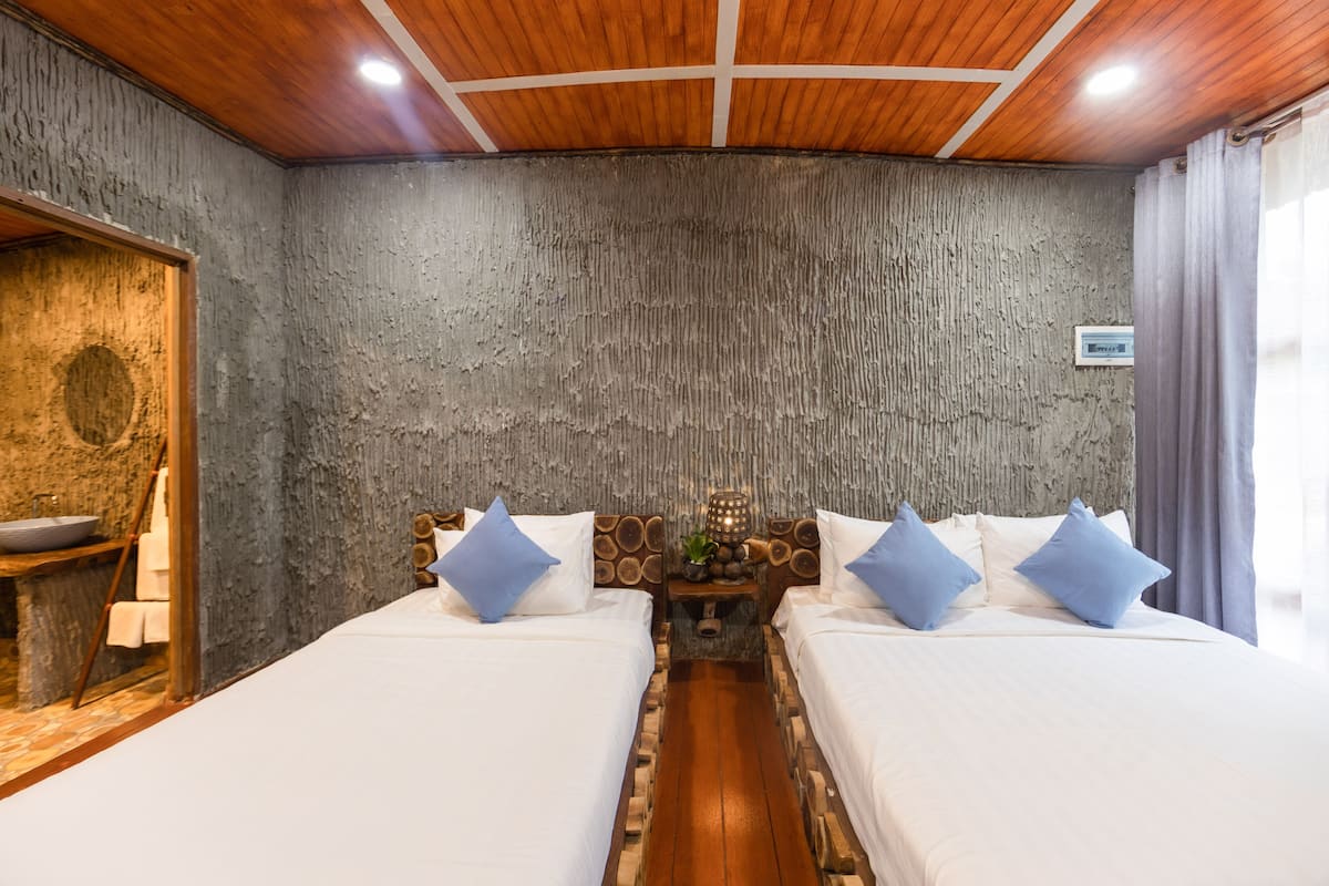 A room with two beds, a wooden ceiling, a textured wall, and a small table with a lamp.