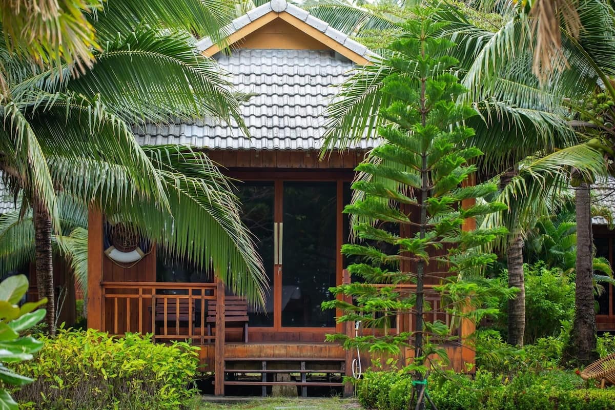 A wooden cabin with a tiled roof, surrounded by lush greenery and palm trees.