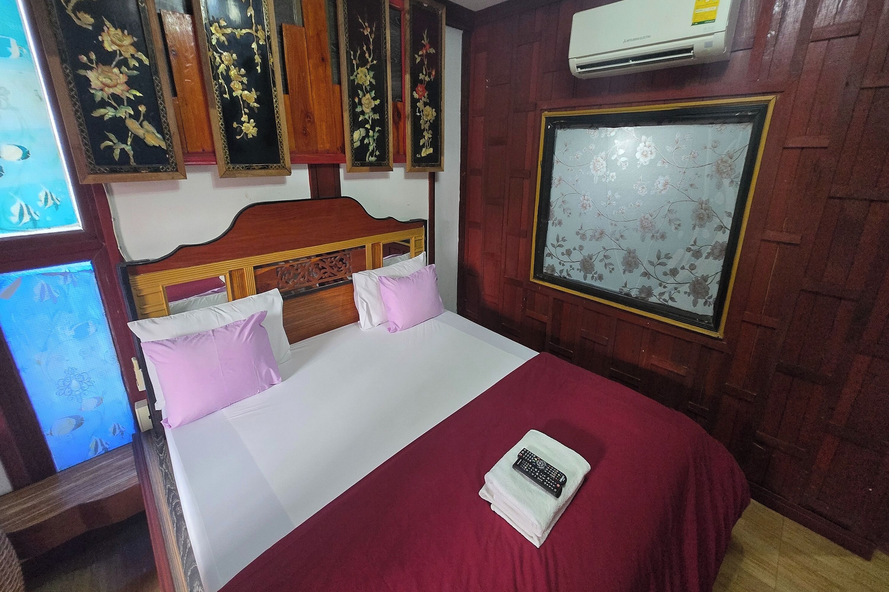 A hotel room with a wooden headboard, a bed with white and pink bedding, a red blanket, and a remote control on a folded towel.
