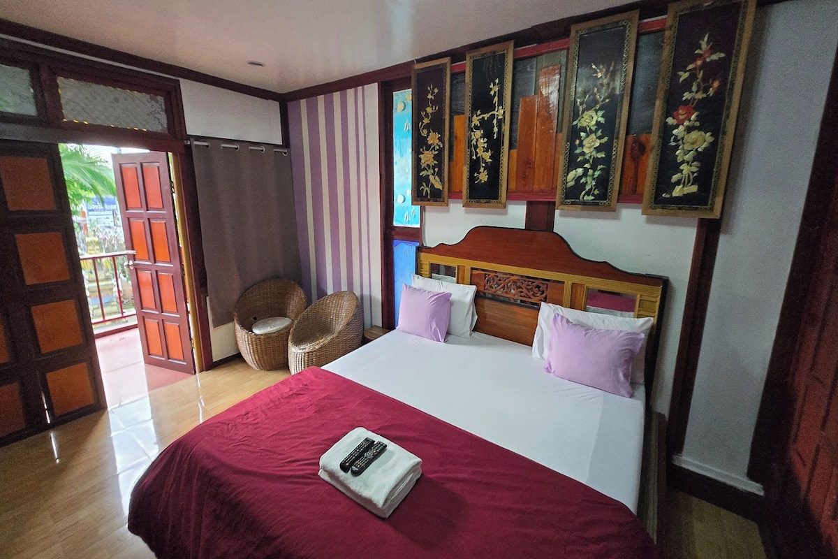 A hotel room with a large bed, a wooden headboard, a red bedspread, and a remote control on the bed. There are wall-mounted floral decorations and a wicker chair in the corner.