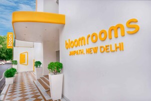 Una entrada de hotel moderna con piso a cuadros, un letrero grande que dice 'bloomrooms Janpath, New Delhi', y un toldo amarillo.
