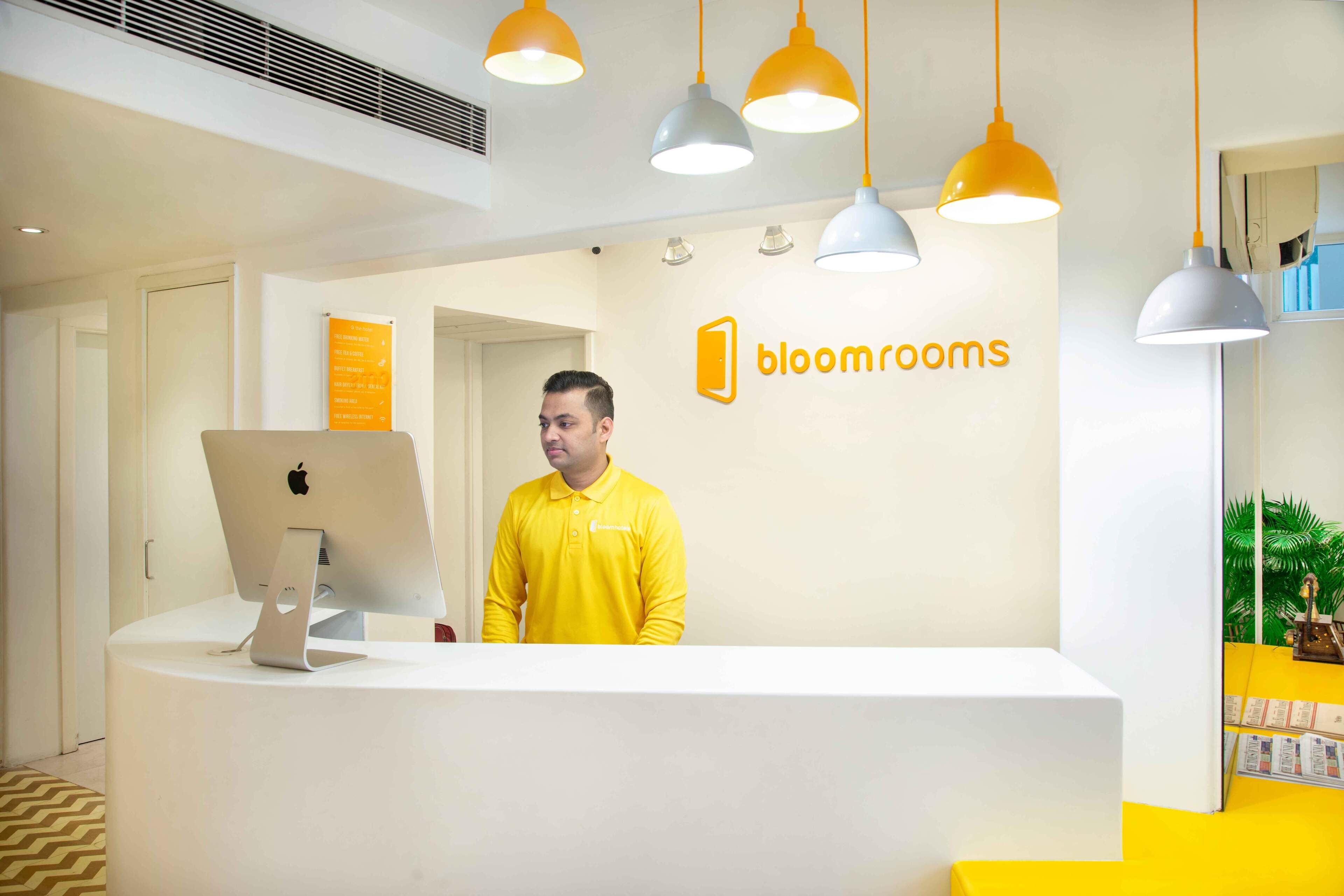 A reception desk with a computer, a person in a yellow shirt, and a wall with the logo "bloomrooms".