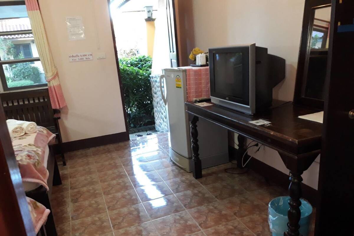 A small kitchenette with a refrigerator, a microwave, and a television. There is a window with curtains, a bed with a rolled-up blanket, and a tiled floor.