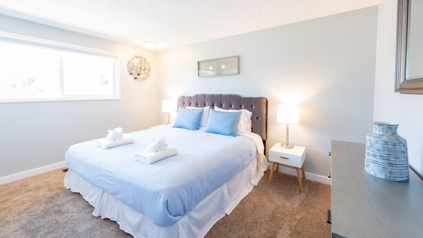A neatly made bed with white and blue bedding, a bedside table with a lamp, and a vase on a dresser. - Comfy & Bright 2BR Flat W/ Fenced Yard Near Washington Square (Tigard)