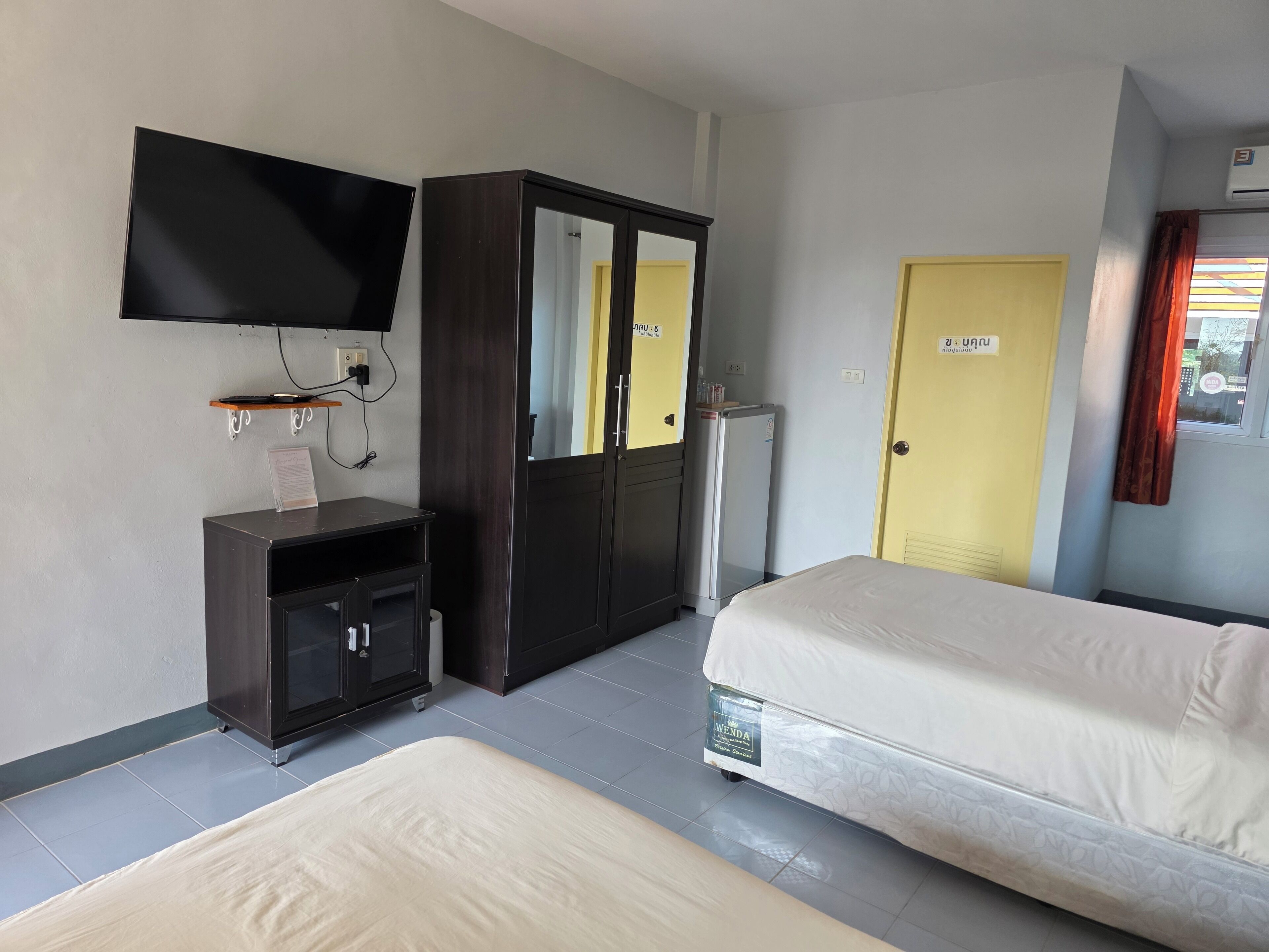 A hotel room with a bed, a TV mounted on the wall, a wardrobe, a small desk with a chair, and a door with a sign.