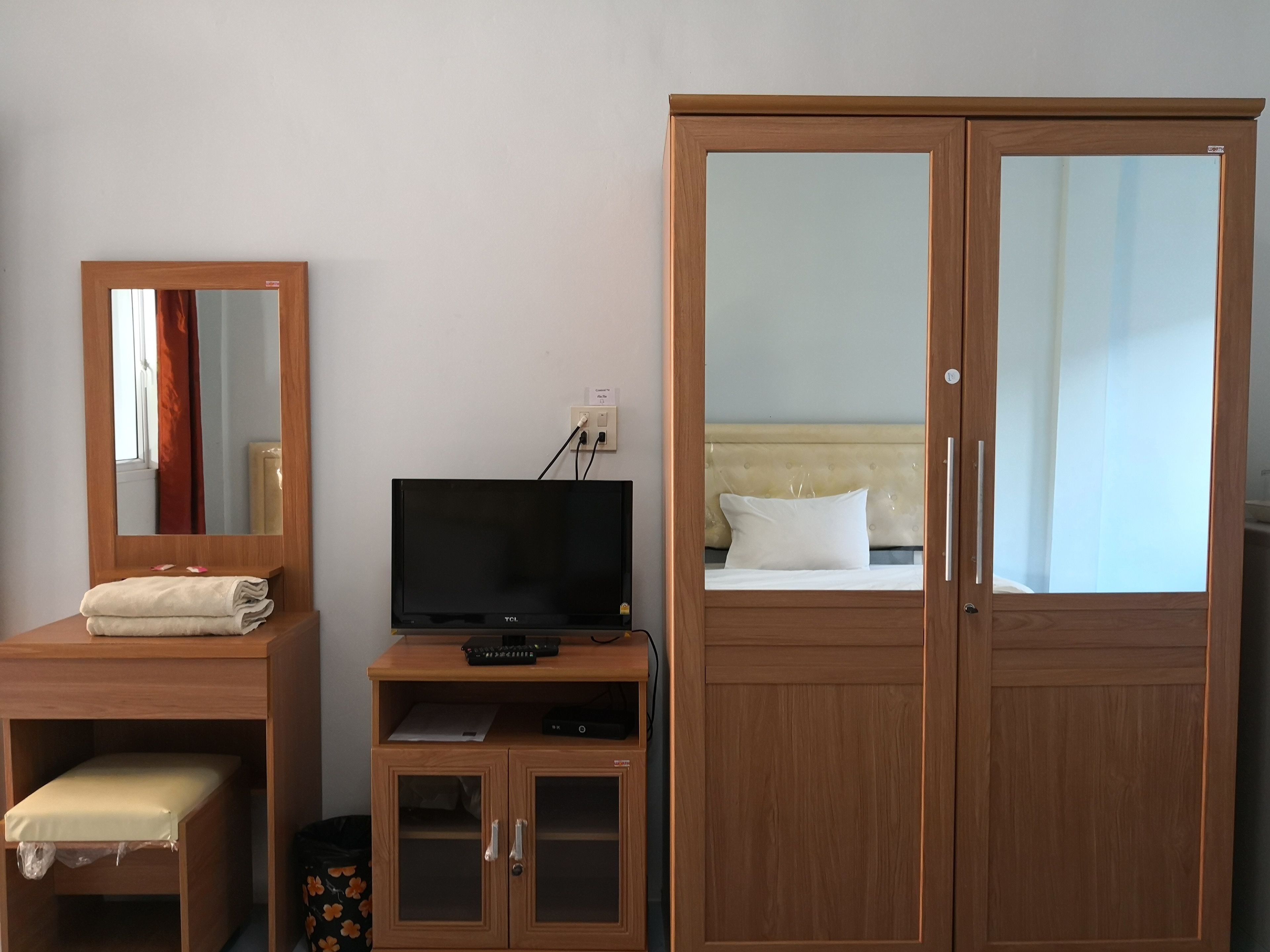 A hotel room with a wooden wardrobe, a television on a wooden stand, a bed with a white pillow, and a mirror on a wooden dresser.