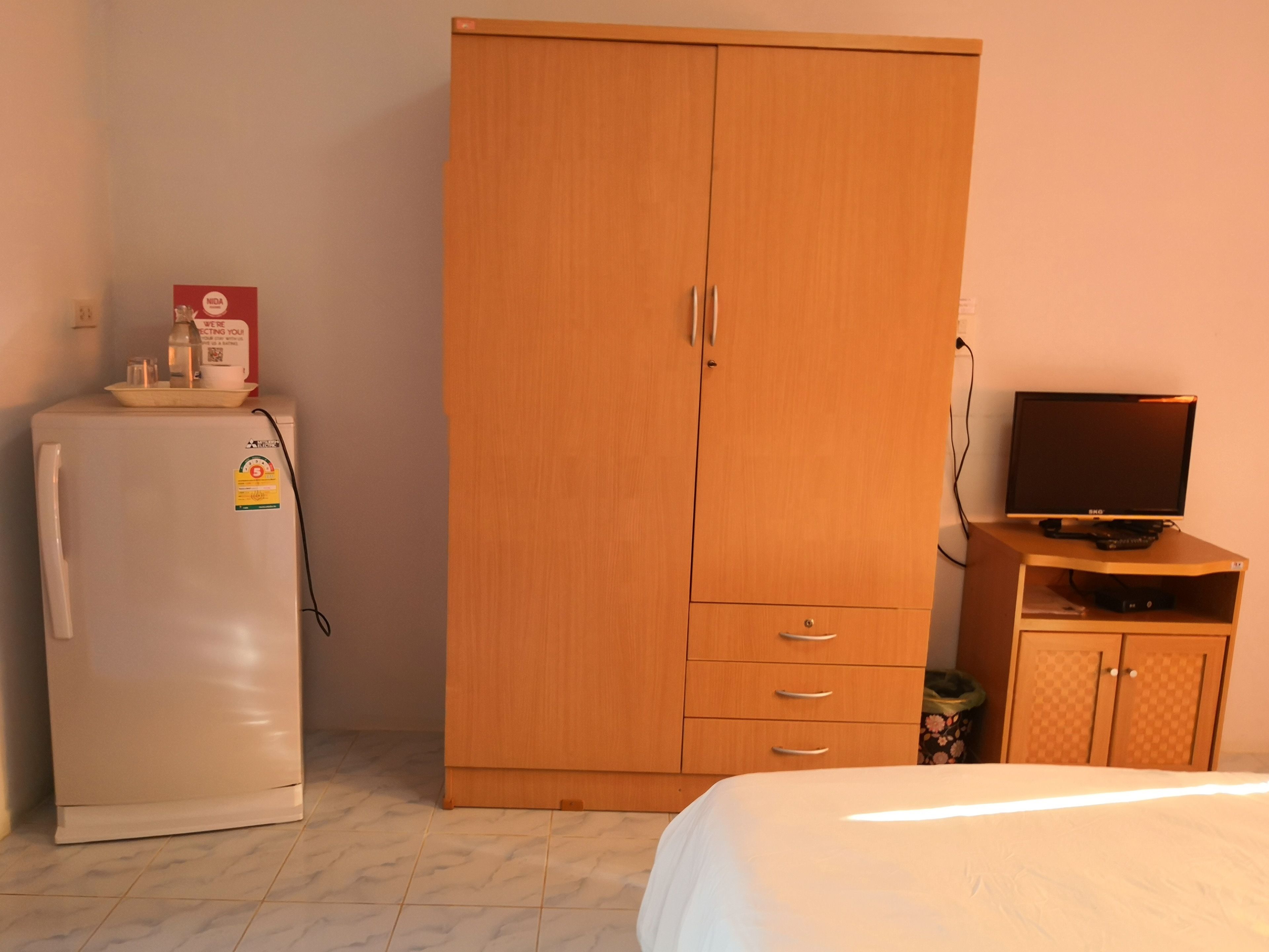 Quarto com guarda-roupa de madeira, frigorífico branco, televisão sobre móvel de madeira e cama com roupa de cama branca.