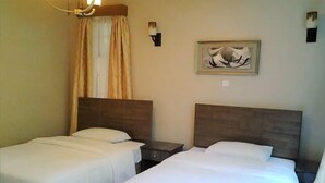 A hotel room with two single beds, a nightstand, a lamp, a painting, and a window with curtains. - Samaritan Homes (Nairobi)