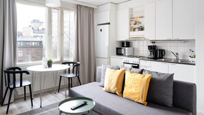 Un espace de vie moderne et compact, comprenant un coin cuisine, un canapé gris agrémenté de coussins jaunes, une table à manger avec des chaises, et une vue sur la ville grâce à de grandes fenêtres.