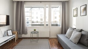 Un salon moderne avec un canapé gris, une petite table ronde ornée d’une plante, une grande fenêtre avec des rideaux et un téléviseur à écran plat posé sur un meuble blanc.