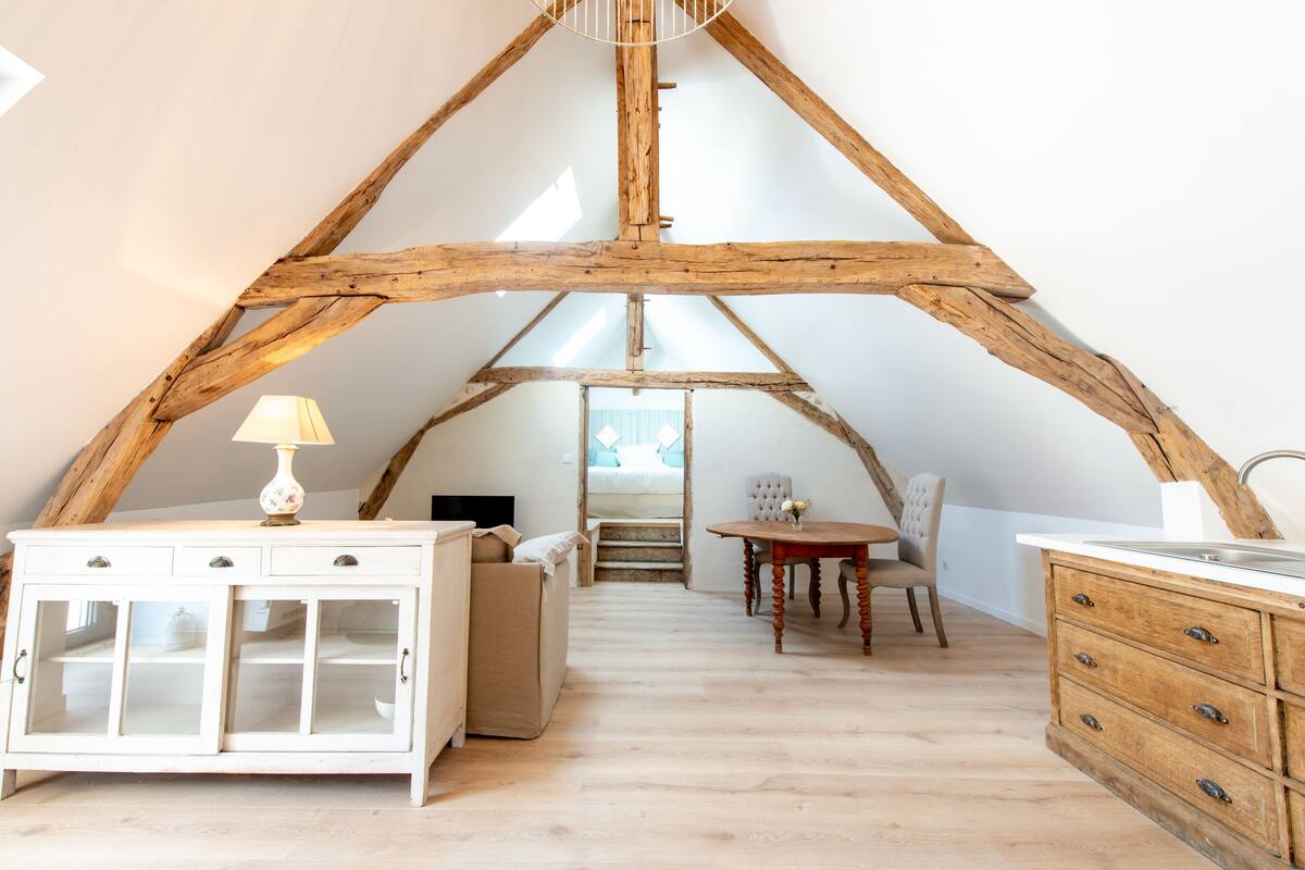 Dormitorio tipo loft con vigas de madera a la vista, un aparador blanco, una mesita de noche con lámpara y vistas a un dormitorio con cama y ventana.
