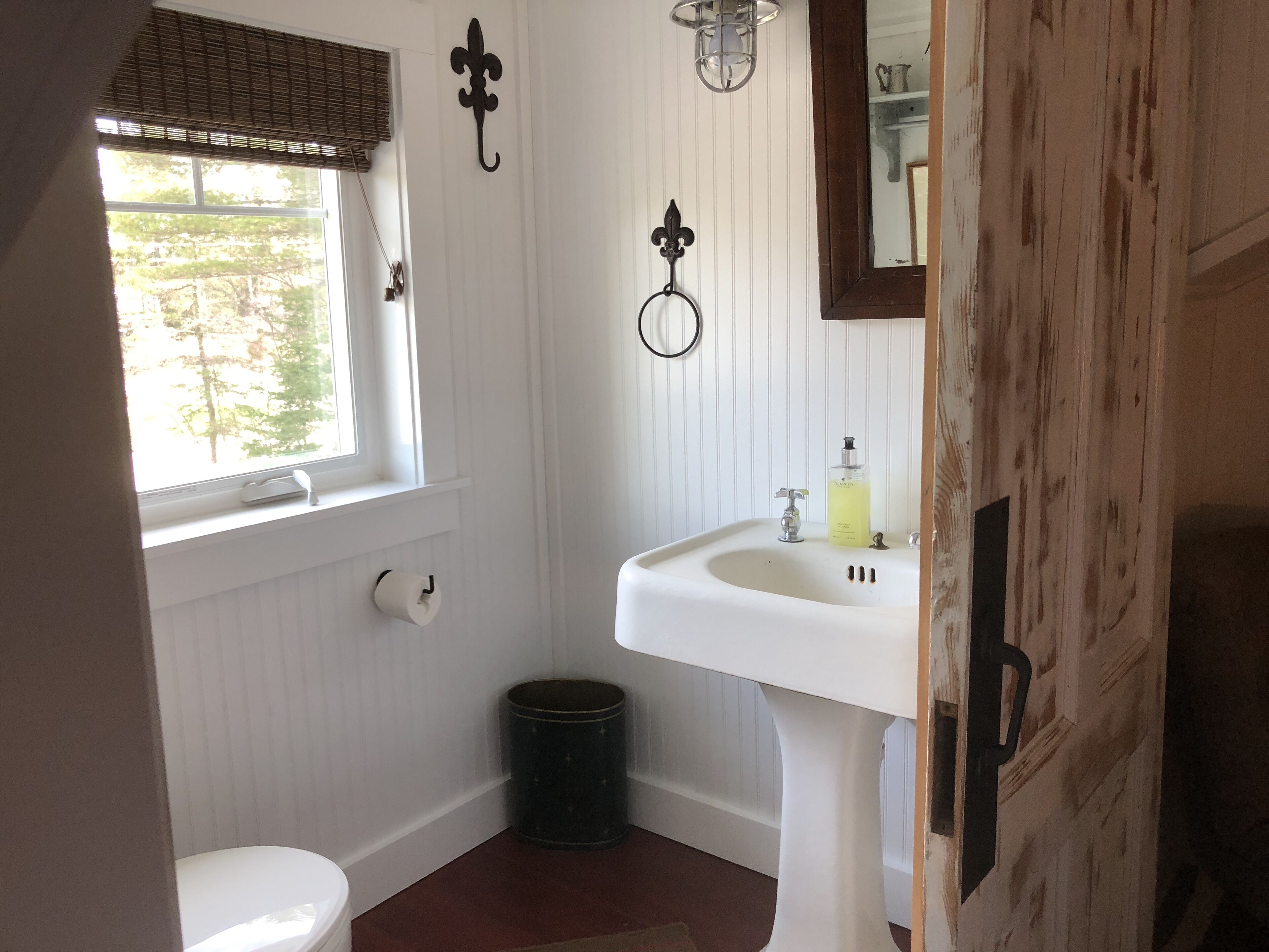 Une petite salle de bain aux murs recouverts de panneaux blancs, équipée de toilettes, d’un lavabo, d’un miroir et d’une fenêtre avec des stores.