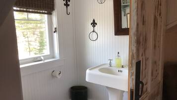 Une petite salle de bain aux murs recouverts de panneaux blancs, équipée de toilettes, d’un lavabo, d’un miroir et d’une fenêtre avec des stores.