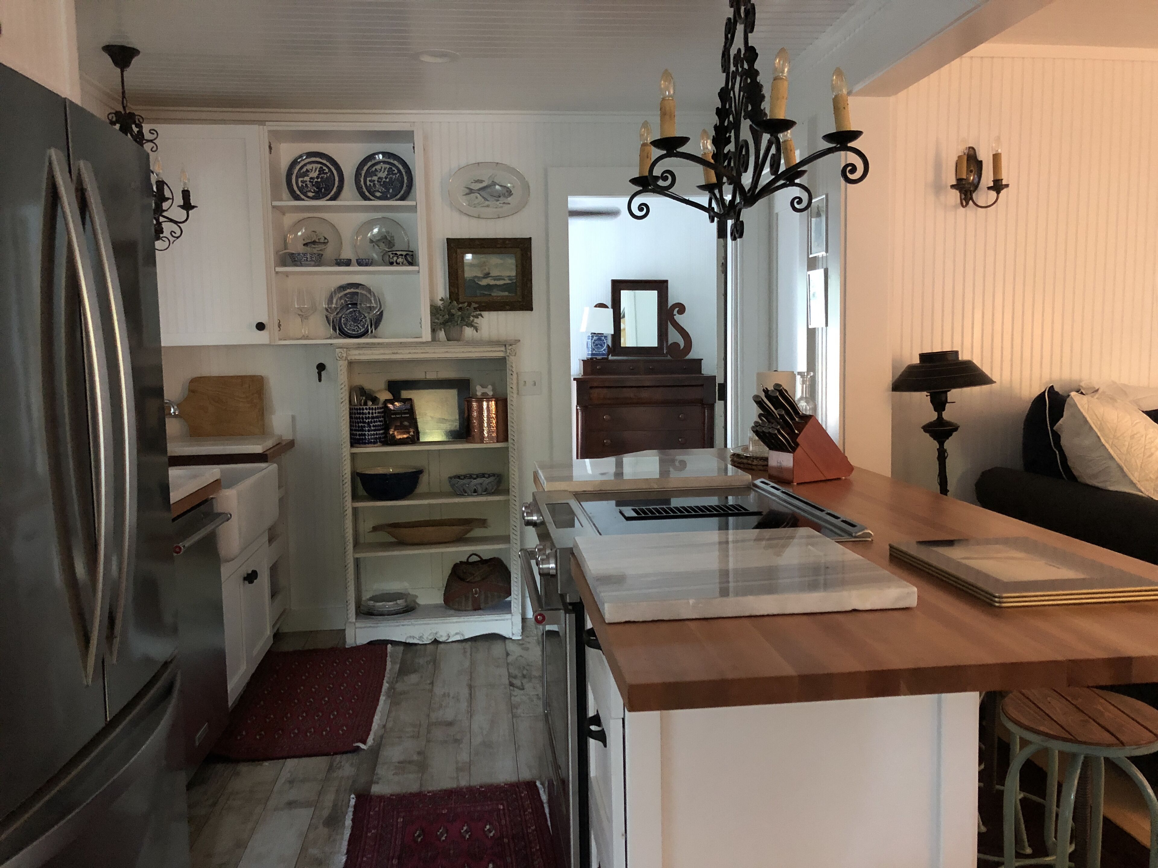 Una cocina con refrigerador, lavamanos y encimera de madera. Hay repisas con vajilla y una araña colgante.