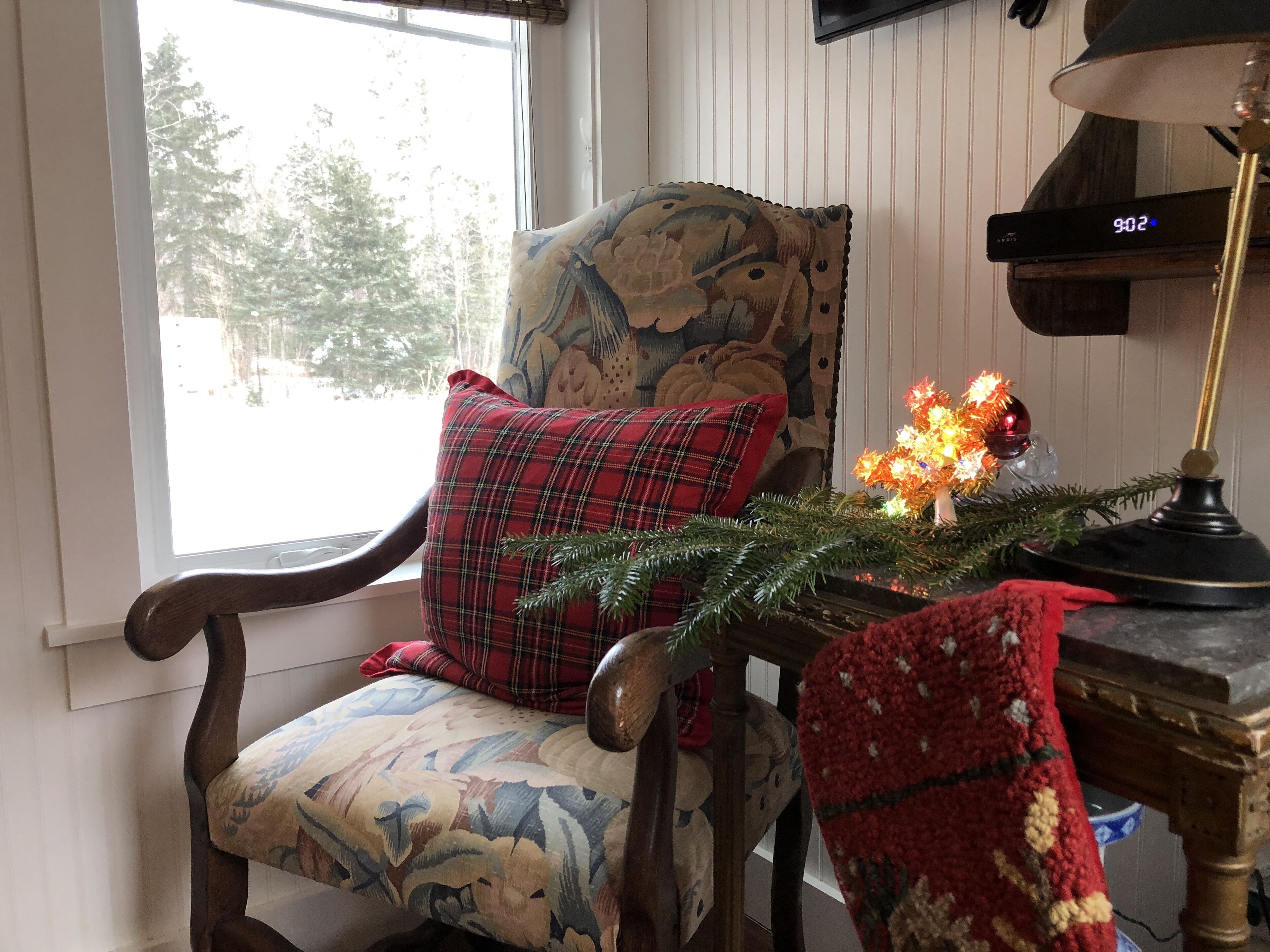 A wooden chair with a floral cushion and a plaid pillow, a small table with a Christmas tree and a lamp, and a wall-mounted clock.