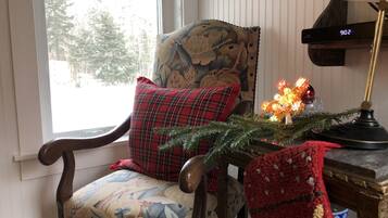 A wooden chair with a floral cushion and a plaid pillow, a small table with a Christmas tree and a lamp, and a wall-mounted clock.