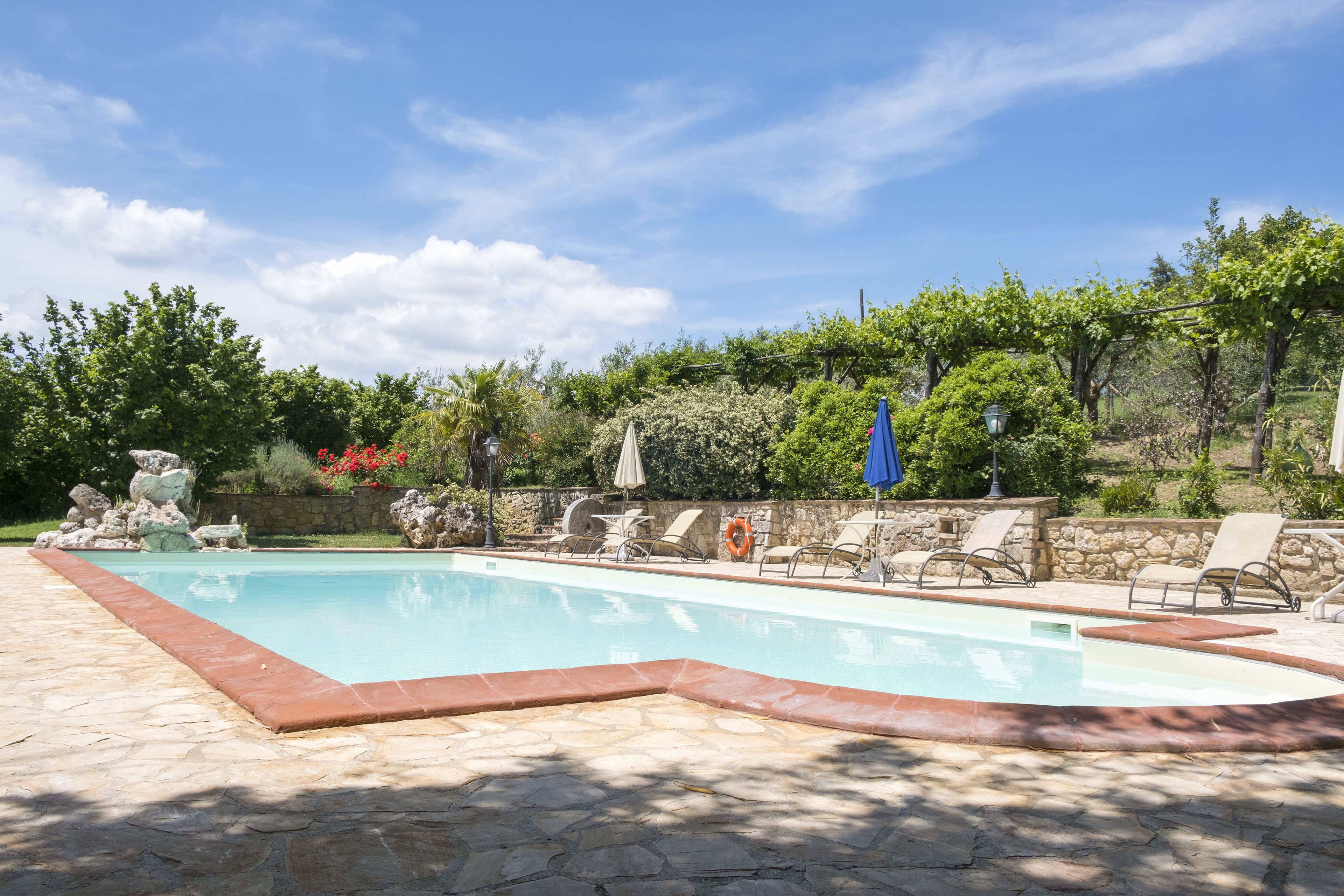 Une piscine aux carreaux de pierre, entourée de chaises longues et de parasols, avec en arrière-plan des arbres et un mur de pierre.