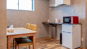 Een kleine keuken met een houten tafel, stoelen, een witte koelkast, een magnetron en een raam.