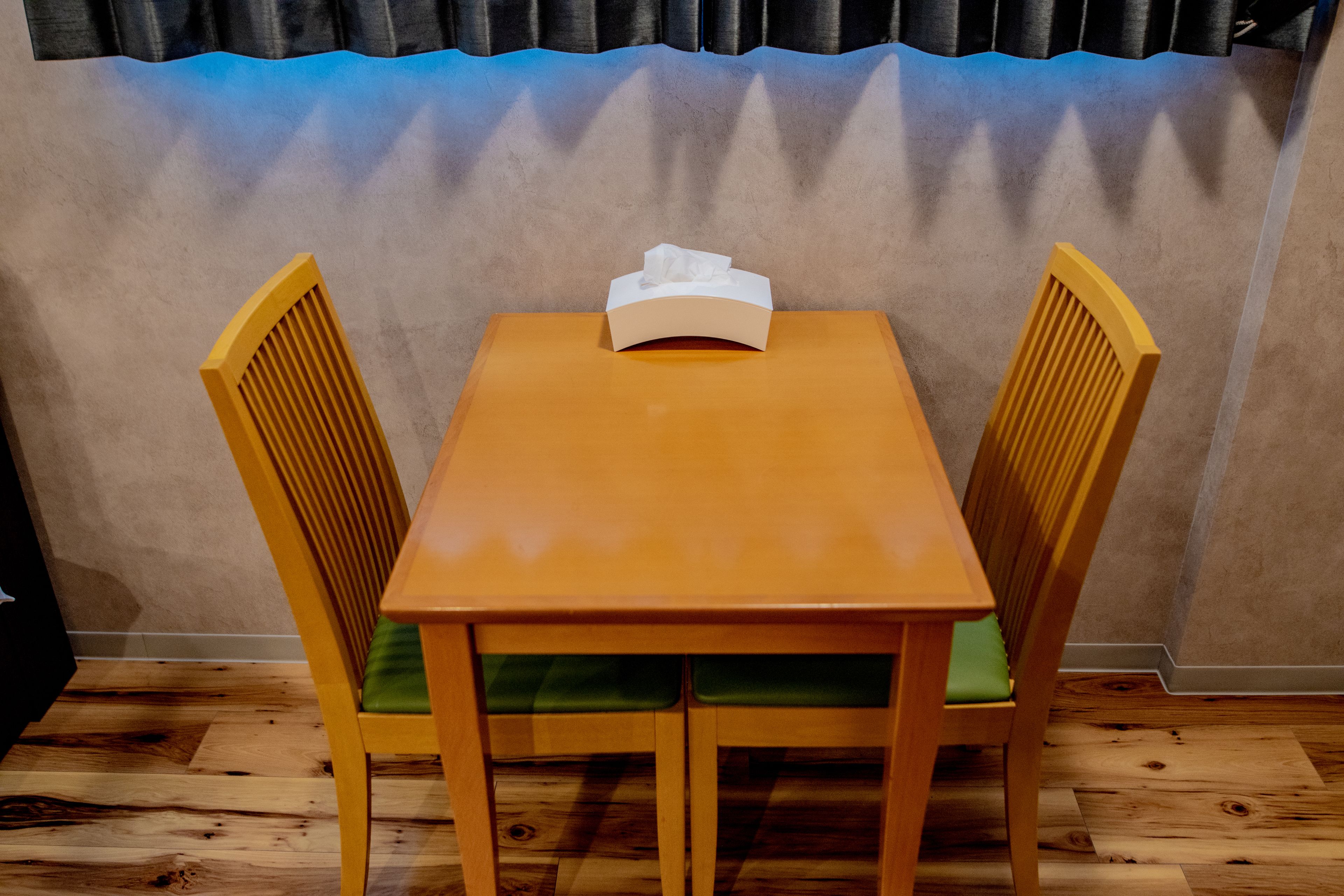Een houten eettafel met een zakvochtige papieren doos erop, omringd door twee houten stoelen met groene kussens.