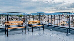 Een balkon met twee houten banken en een metalen hekwerk, met uitzicht op een stadsgezicht met gebouwen en bergen in de verte.
