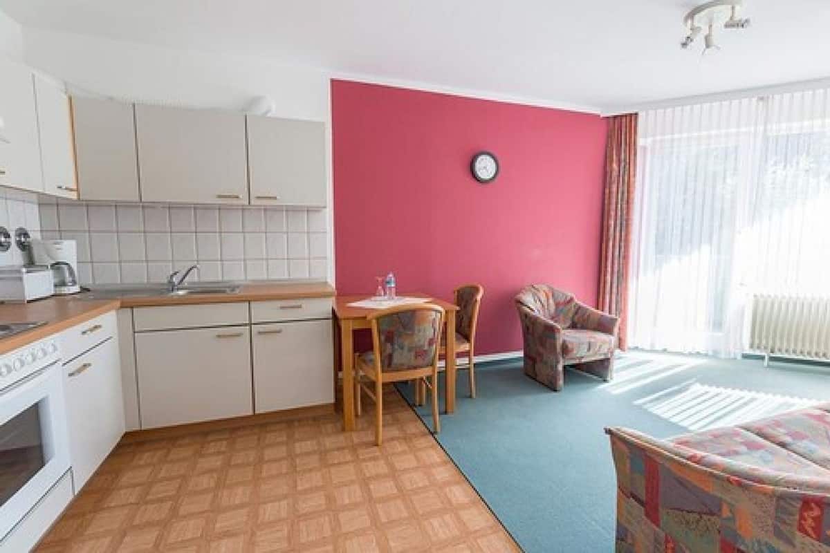 A compact kitchen with white cabinets, a wooden countertop, and a dining area with two chairs. A clock is mounted on the pink wall, and a radiator is visible in the corner.