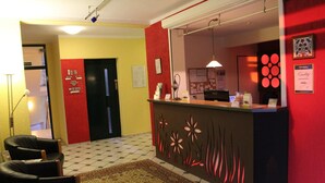 Zona de recepción con mostrador de entrada, pared roja, alfombra estampada y puerta con indicaciones.