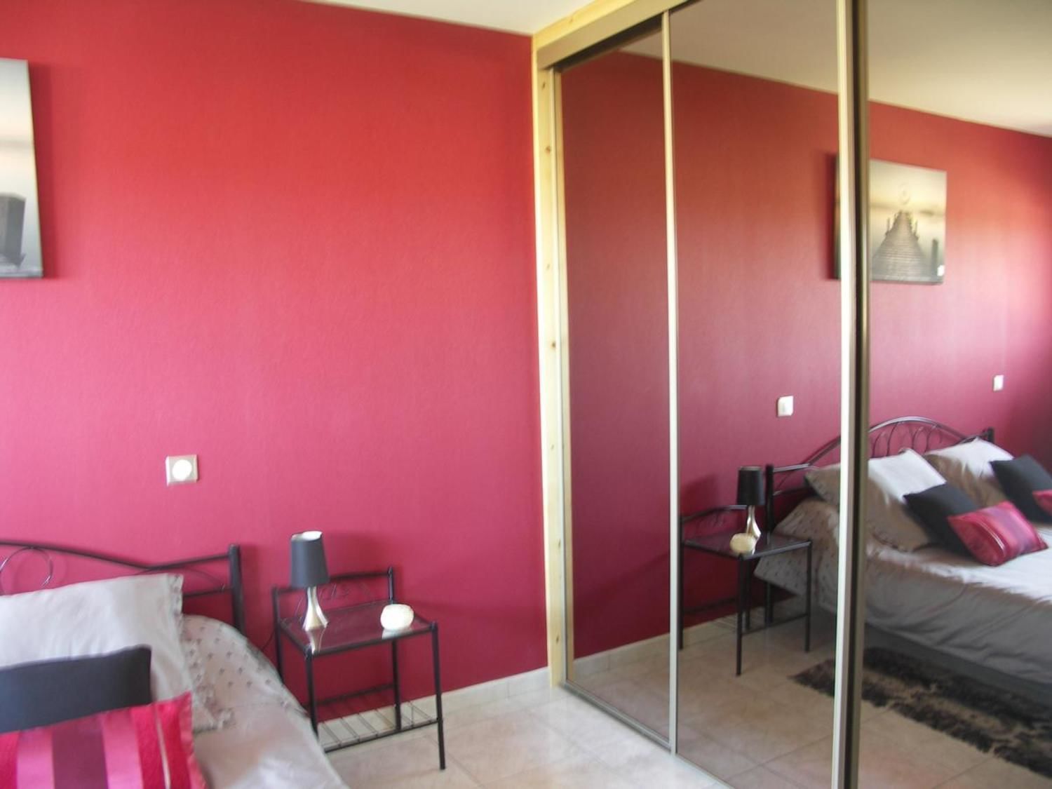 Ein Schlafzimmer mit roter Wand, einem Bett mit schwarzem Gestell, einem kleinen Tisch mit Lampe und einer Glastür, die in einen anderen Raum führt.