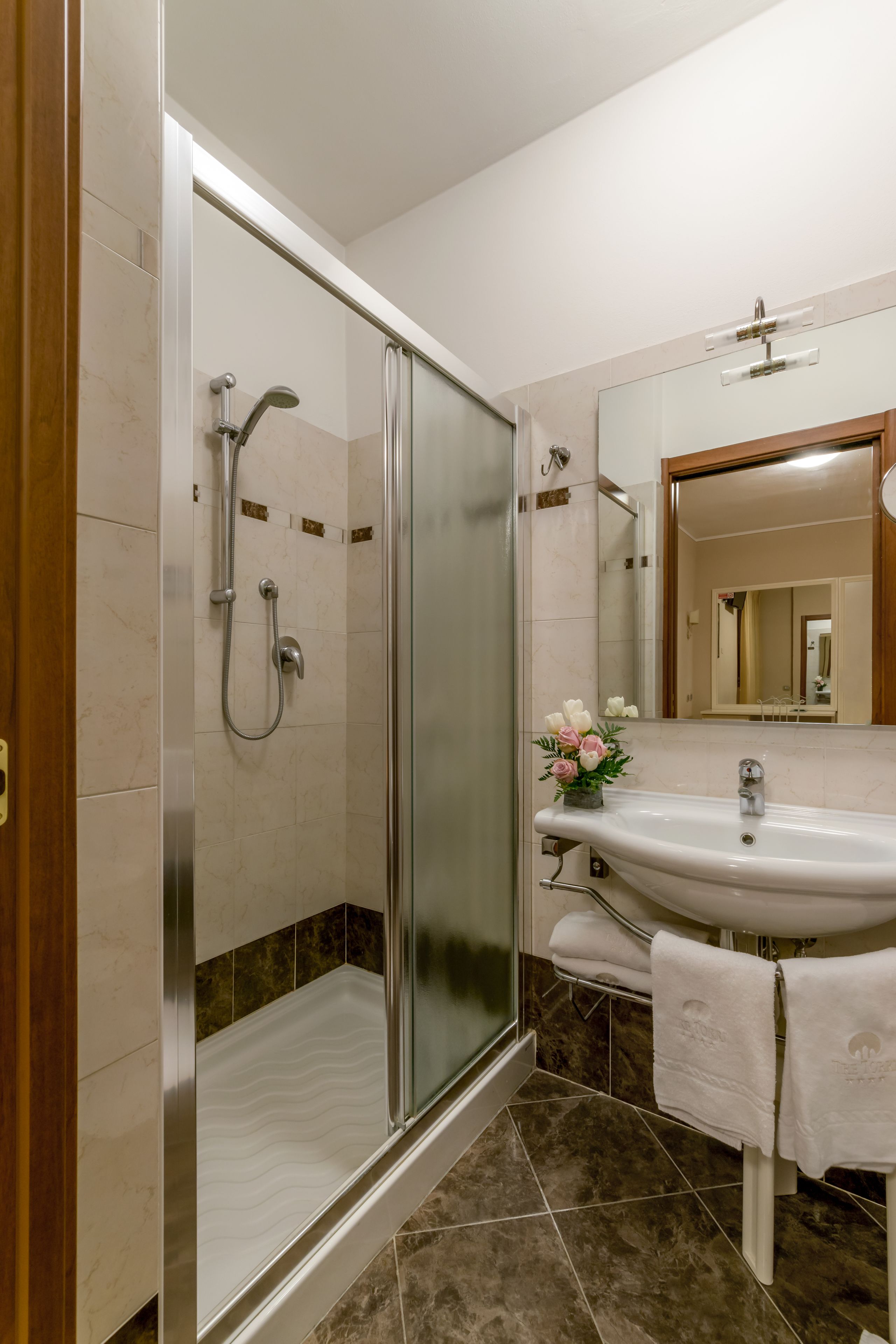 Une salle de bain moderne avec une douche à parois de verre, un lavabo blanc avec un miroir au-dessus, et un petit arrangement floral posé sur le lavabo.