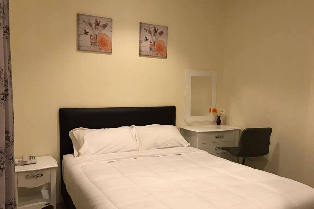 A neatly made bed with white linens, a black headboard, a bedside table with a lamp, a chair, and two wall-mounted paintings.