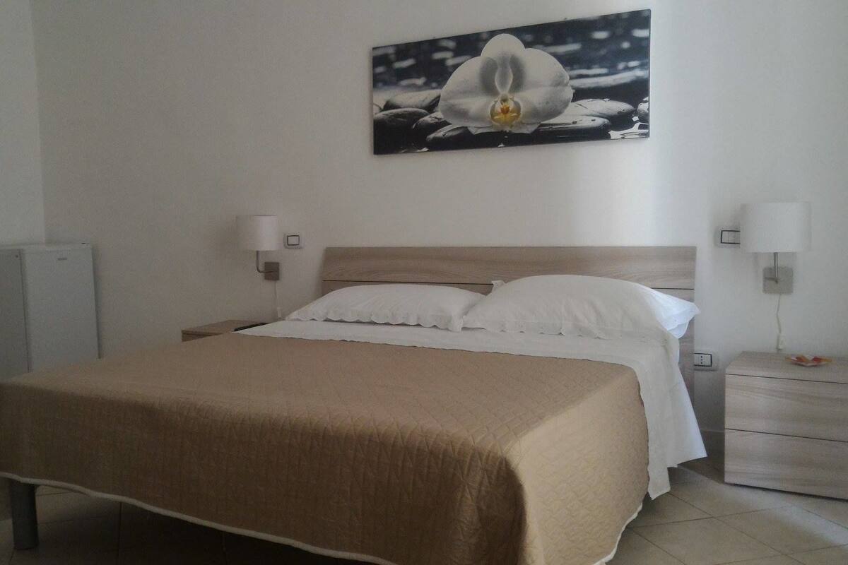 A neatly made bed with a beige blanket, a wooden headboard, and white pillows. There is a wall-mounted picture of a flower, and two bedside lamps on either side of the bed.