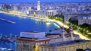 Paesaggio costiero al crepuscolo con una passeggiata sul lungomare ben visibile, edifici storici e una grande struttura a cupola.