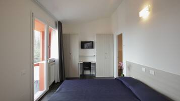 Een slaapkamer met een bed, een bureau, een stoel, een televisie en een schuifdeur naar een balkon.