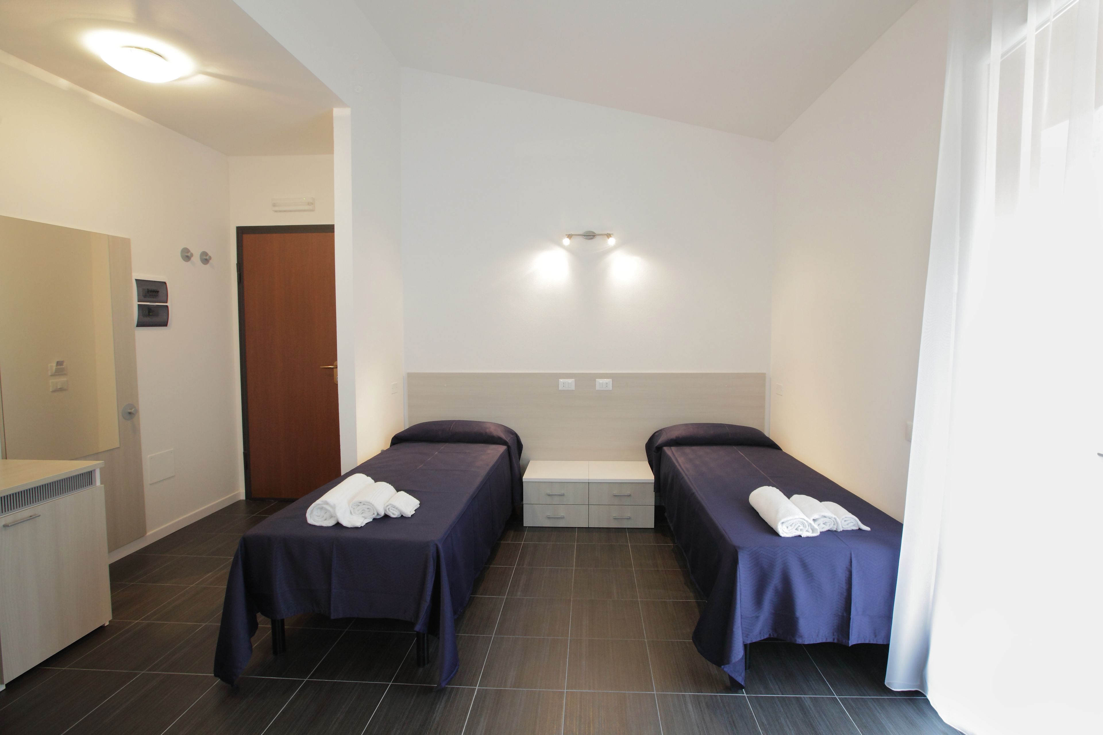 A hotel room with two beds, a small table with towels, a wooden door, and a white wall with a light fixture.