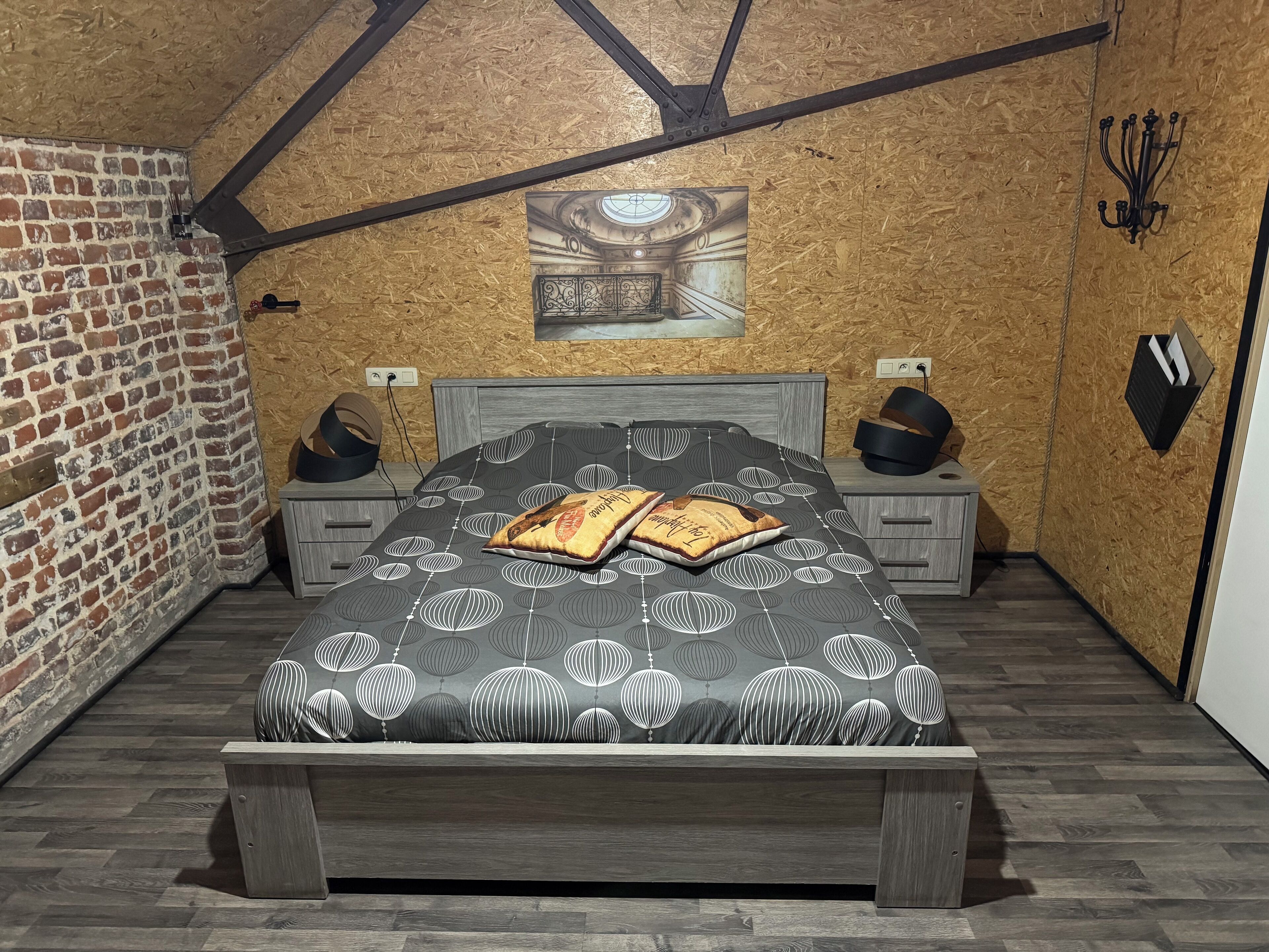 Une chambre avec un mur en briques, un plafond en bois, un lit avec une literie à motifs, deux tables de chevet avec des lampes et un luminaire fixé au mur.