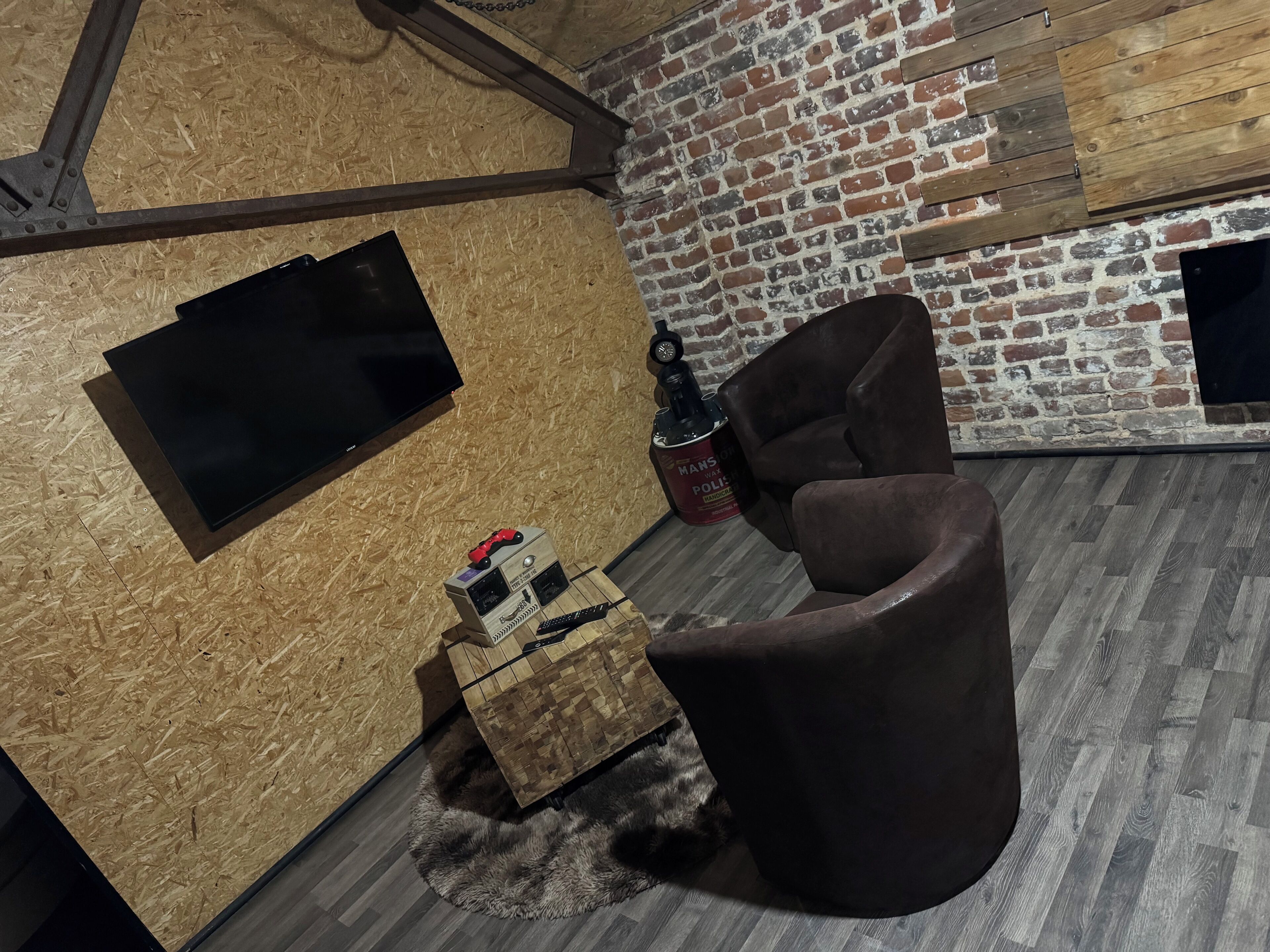 Una habitación acogedora con un televisor instalado en una pared de madera, un sillón marrón, un pequeño cofre de madera y una pared de ladrillo.