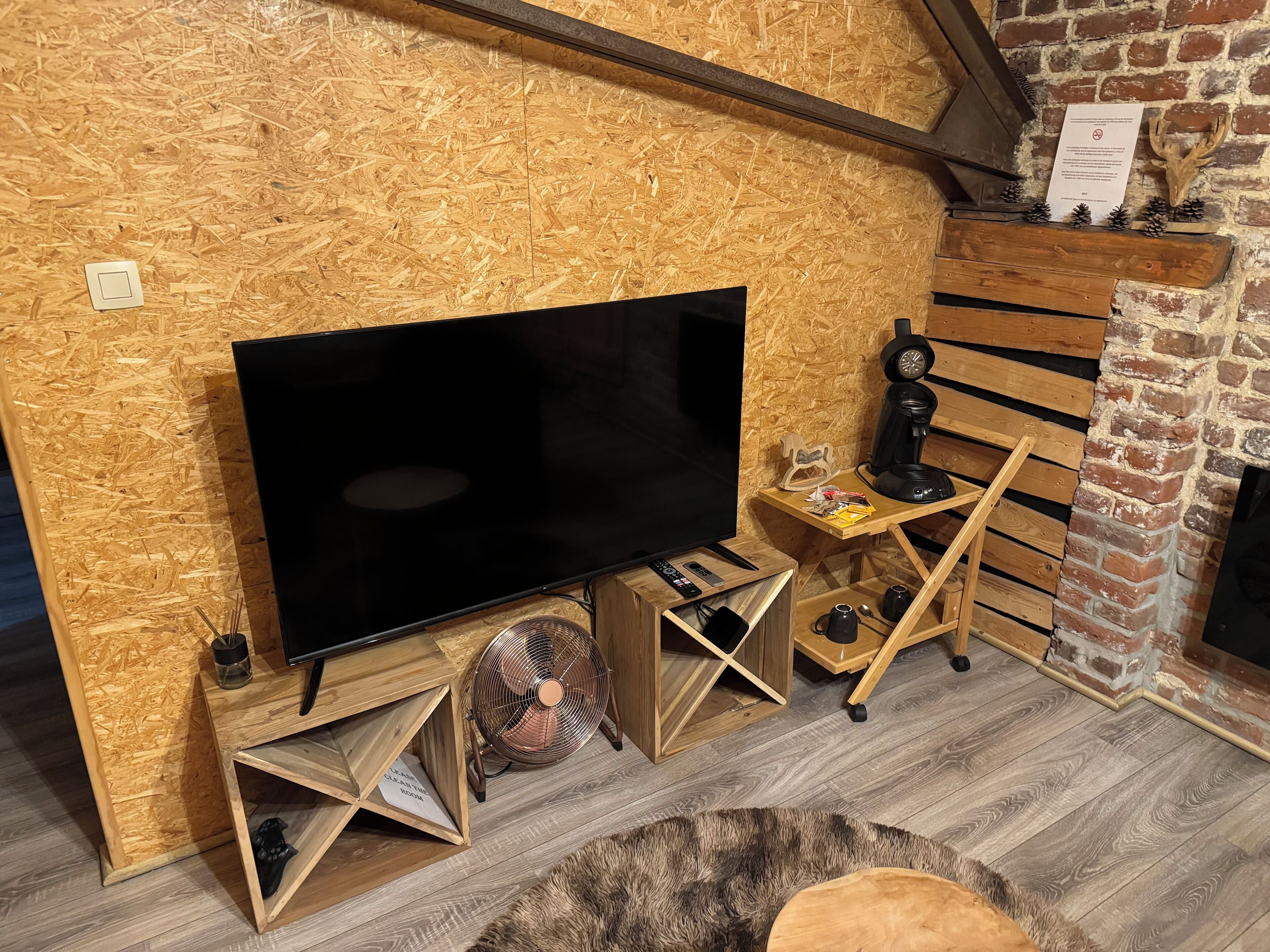 A cozy room with a large flat-screen TV on a wooden stand, a small wooden table with a coffee maker, and a fireplace with a brick wall.