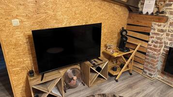 A cozy room with a large flat-screen TV on a wooden stand, a small wooden table with a coffee maker, and a fireplace with a brick wall.