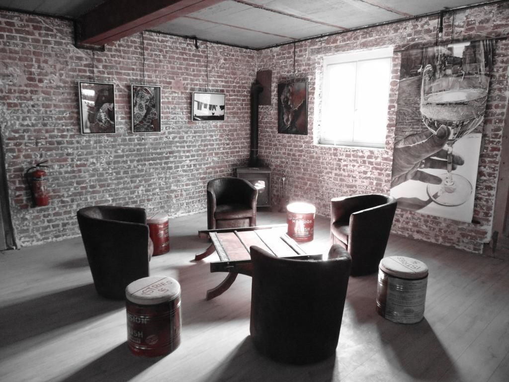 A rustic room with exposed brick walls, a wooden table, and several black chairs. There are cans on the floor, a fire extinguisher on the wall, and a large window.